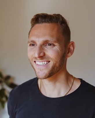 Steven Celi smiling in a black t-shirt, in a bright room with a plant